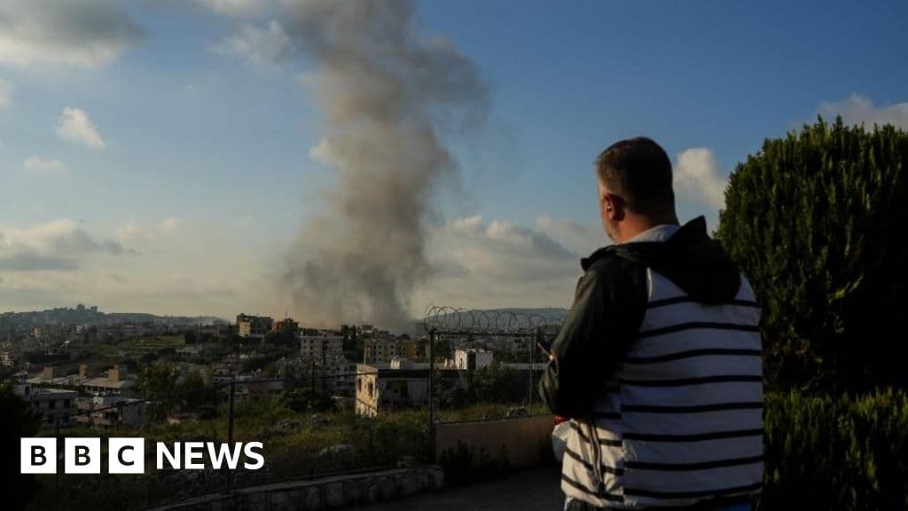 A man in a grey and black striped jacket looks at smoke rising from a building following an Israeli airstrike, he is holding a smart phone.